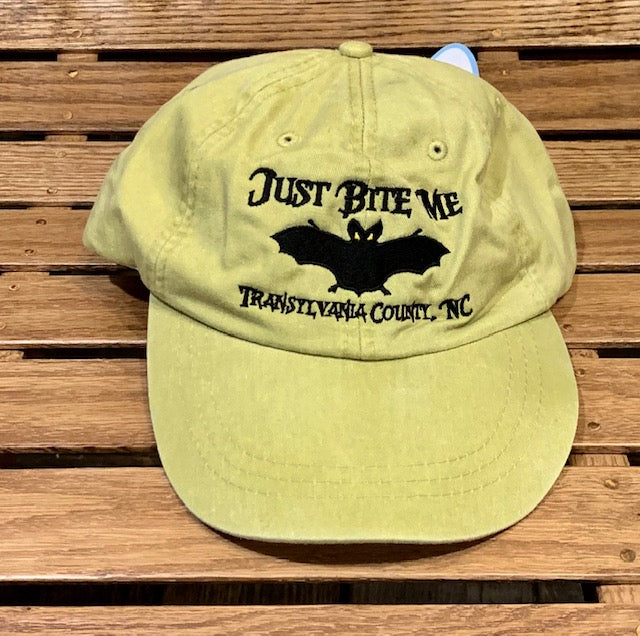 A khaki yellow baseball cap with 'Just Bite Me' embroidered on the front and a bat silhouette and Transylvania County, NC.