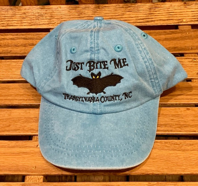 A sky blue baseball cap with 'Just Bite Me' embroidered on the front and a bat silhouette and Transylvania County, NC.