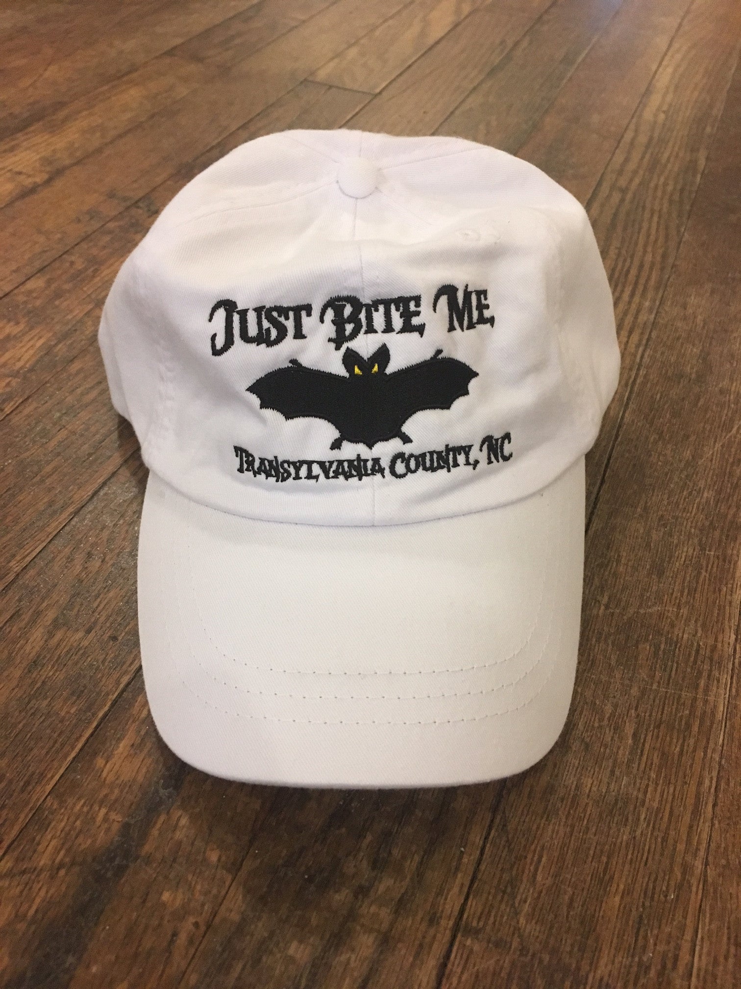 A white baseball cap with 'Just Bite Me' embroidered on the front and a bat silhouette and Transylvania County, NC.