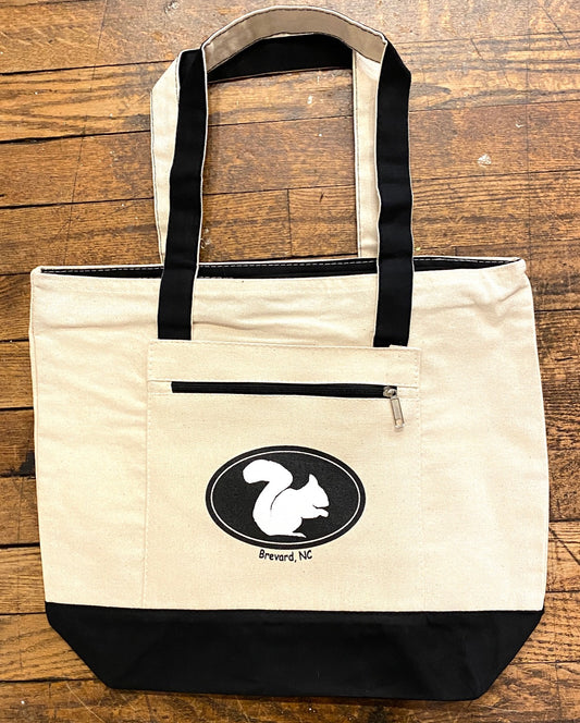 A beige canvas tote bag with a white squirrel design and the text 'Brevard, North Carolina'. It has a black front pocket with a zipper and black straps.