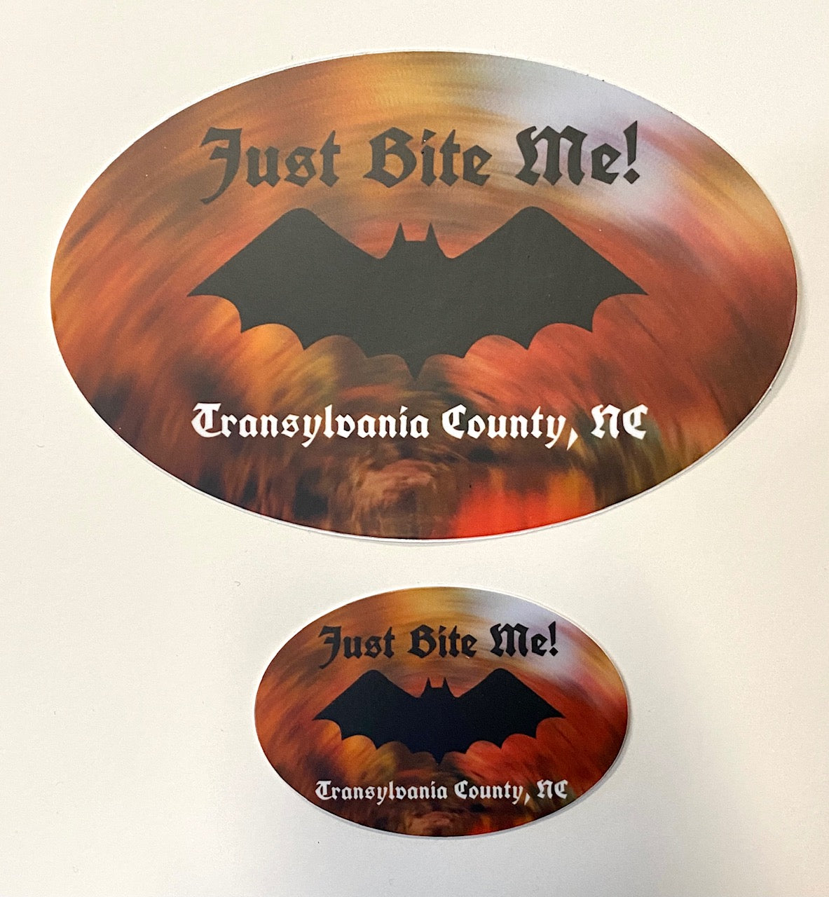Two oval decals with a cosmic background featuring a black bat and the text 'Just Bite Me, Transylvania County, NC'. They come in two sizes displayed next to each other.