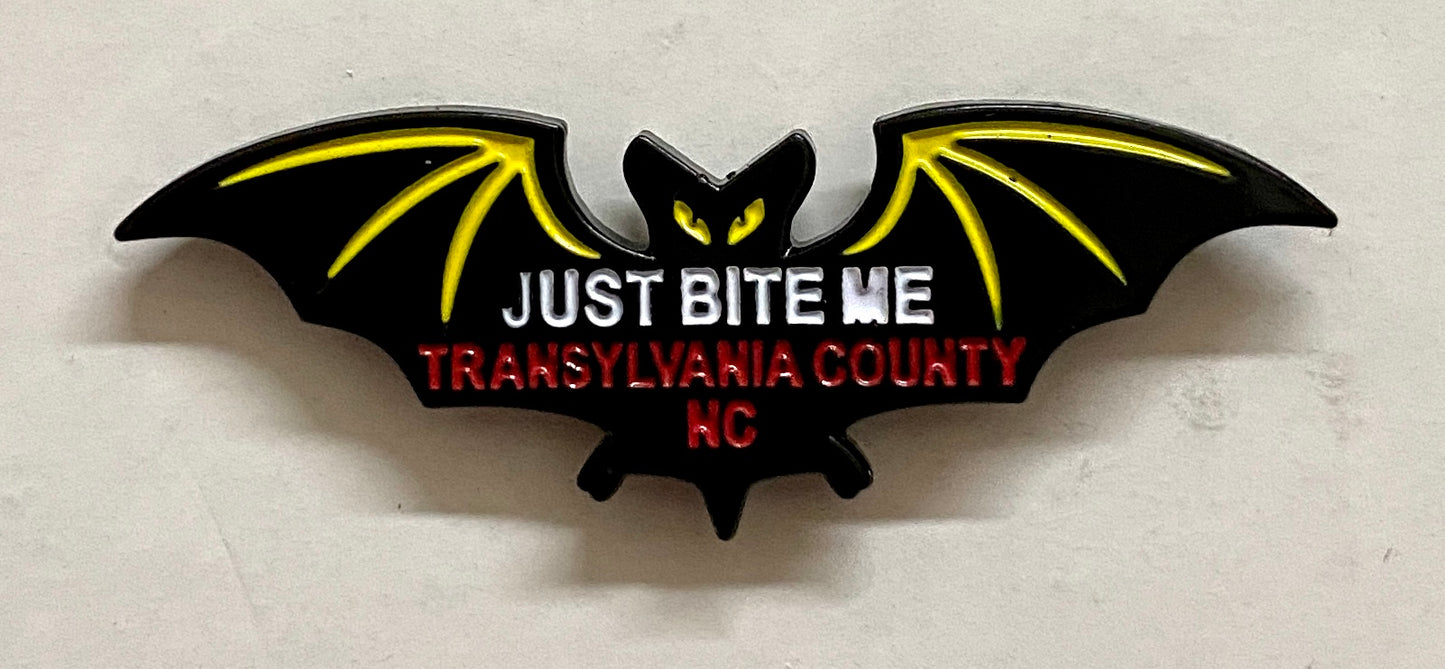 A metal black bat hat/lapel pin with the text 'JUST BITE ME, TRANSYLVANIA COUNTY, NC' on it.