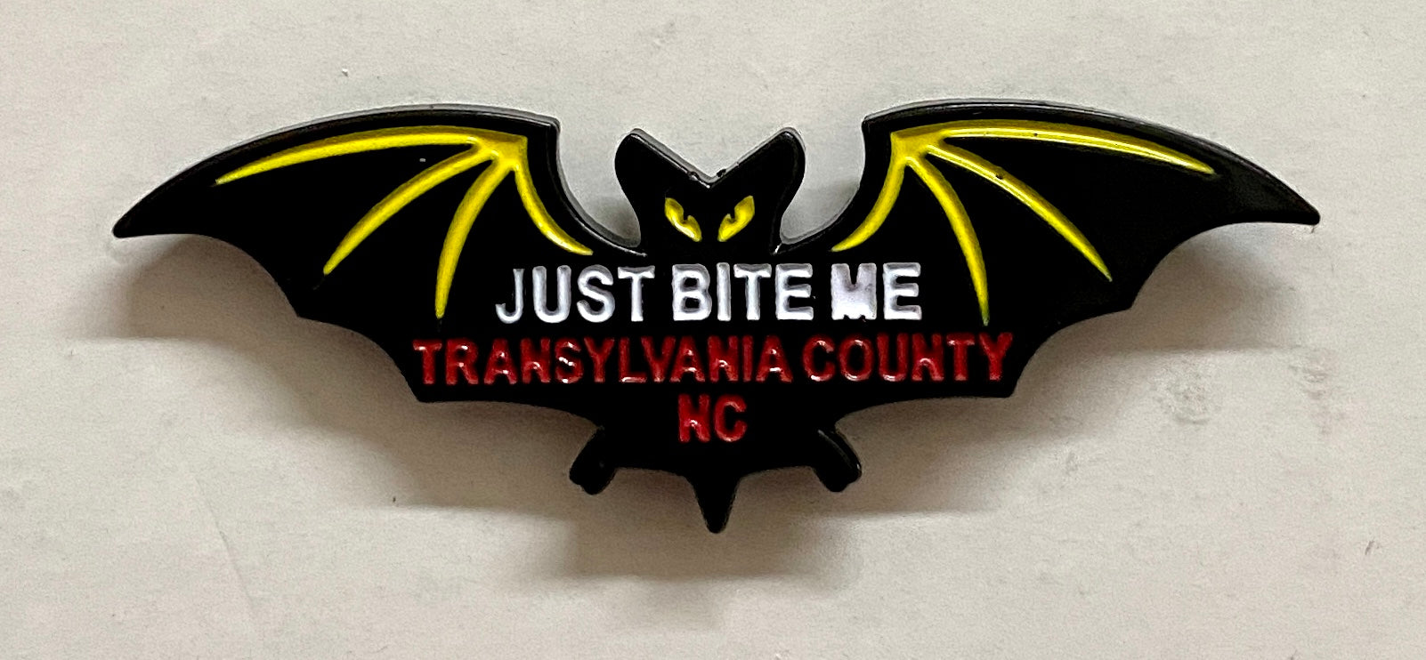 A metal black bat hat/lapel pin with the text 'JUST BITE ME, TRANSYLVANIA COUNTY, NC' on it.