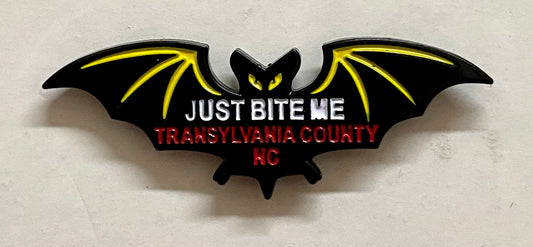 A metal black bat hat/lapel pin with the text 'JUST BITE ME, TRANSYLVANIA COUNTY, NC' on it.