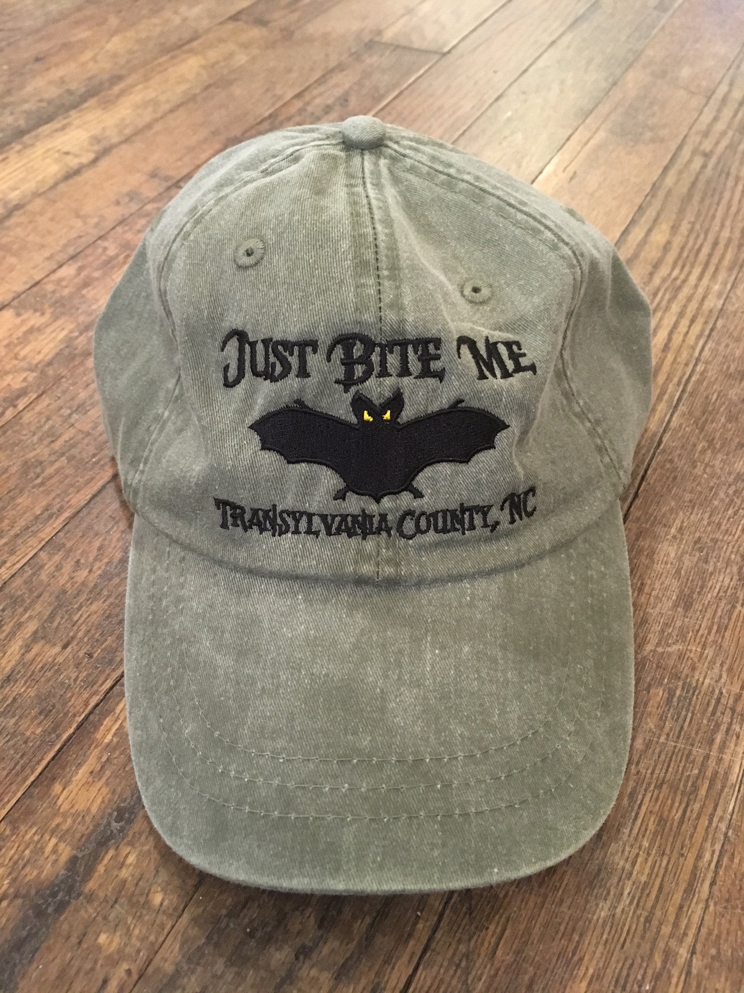 A green baseball cap with 'Just Bite Me' embroidered on the front and a bat silhouette, indicating a location connection to Transylvania County, NC.