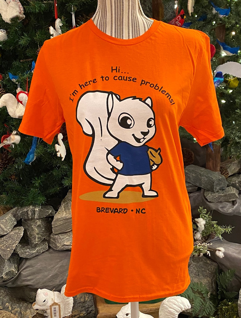 An orange short-sleeved crew neck t-shirt with a graphic of a white squirrel holding a beer and the phrase 'Hi, I'm here to cause problems.