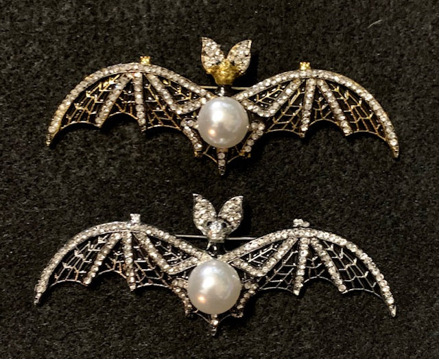 A brooch designed as a bat with extended wings, featuring a central pearl and intricate detailing. Choose silver or gold.