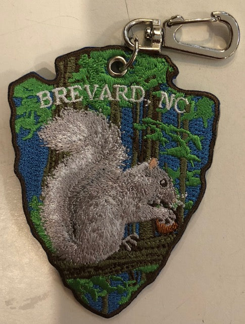 An embroidered key clip in the shape of an arrowhead with 'Brevard, NC' and a white squirrel eating a nut on it.