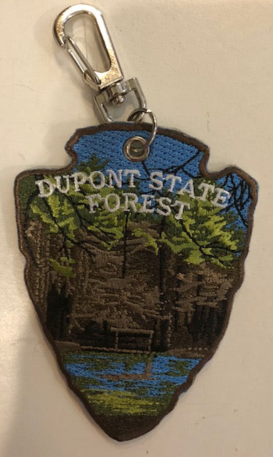 A key chain with an embroidered design featuring the words 'DUPONT STATE FOREST' on a background forest scene in the shape of an arrowhead.