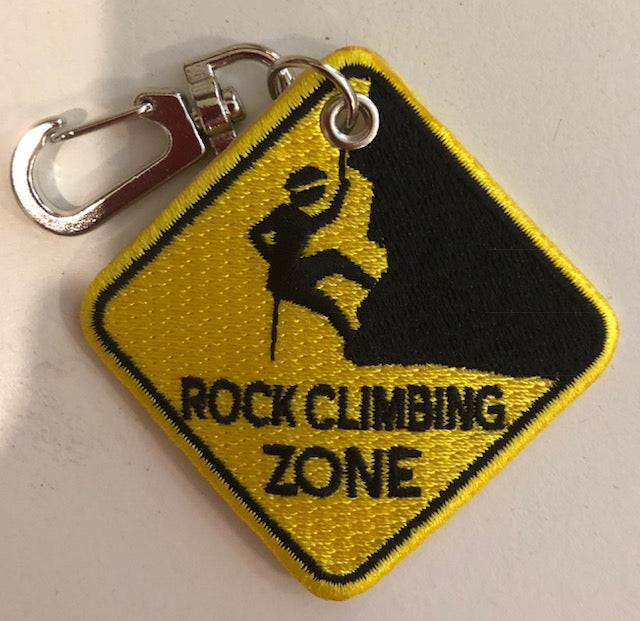 A yellow and black embroidered key clip with 'ROCK CLIMBING ZONE' text and a silhouette of a person climbing on one side.