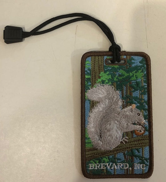 Embroidered luggage tag featuring a white squirrel with 'Brevard, NC' text, attached to a black strap.