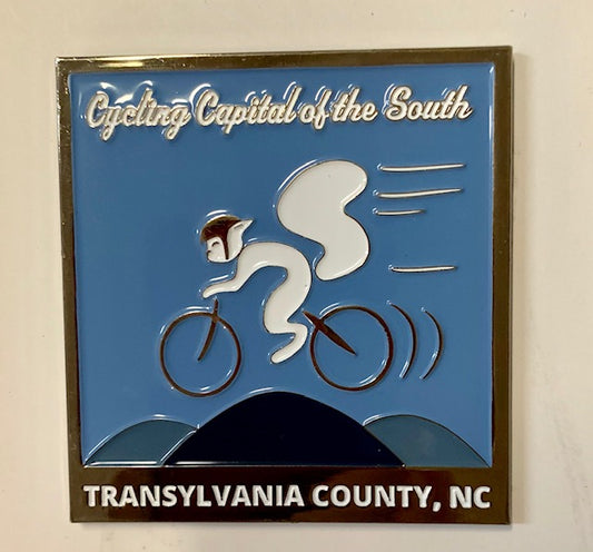 A square metal magnet with an illustration of a white squirrel cyclist and the text 'Cycling Capital of the South, Transylvania County, NC'.