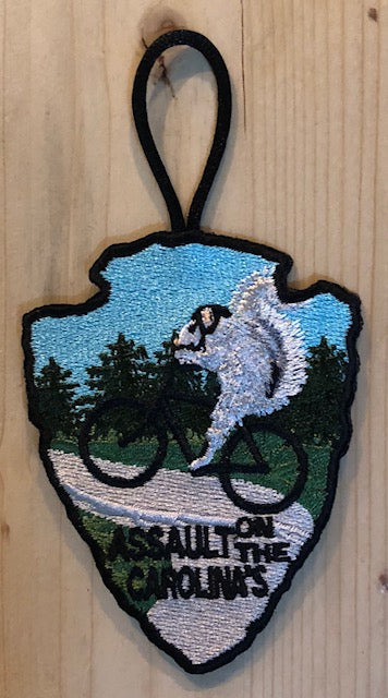 An embroidered ornament depicting a white squirrel riding a bicycle with Assault on the Carolina's embroidered.