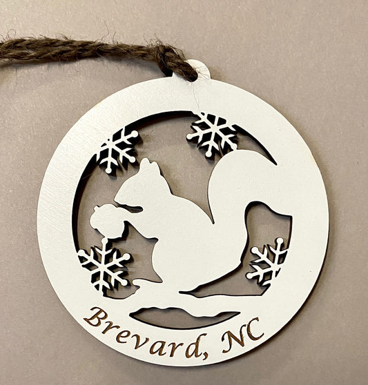 A laser cut wood ornament in the shape of a circle with a white squirrel and snowflakes design, and the text 'Brevard, NC'.