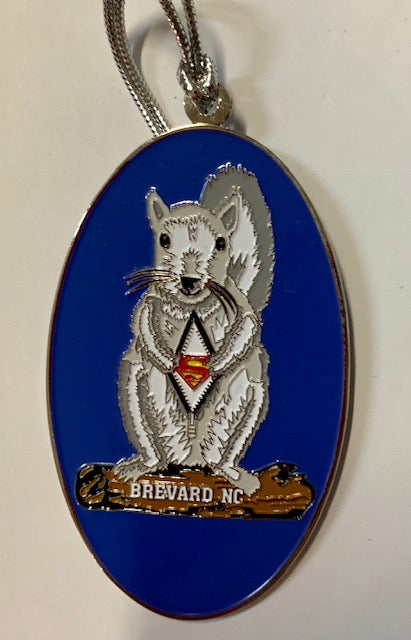 A metal ornament with a white squirrel with a Superman emblem on its chest. Blue background, with Brevard, NC on the log the squirrel is standing on.
