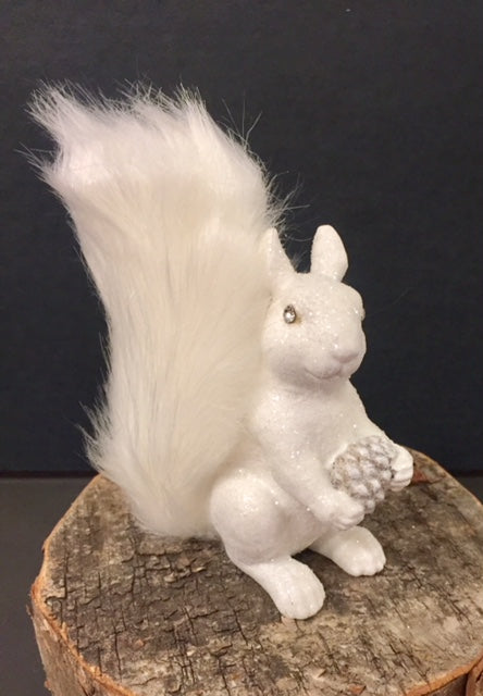 A white, sparkly squirrel Christmas ornament with fluffy tail and rhinestone eyes, holding a pine cone. Without loop to hang.