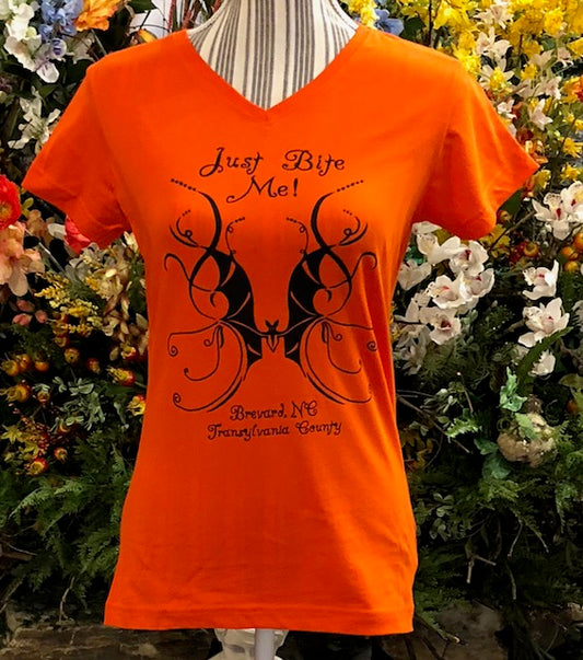 An orange short sleeve V-neck women's t-shirt with a swirly bat design and the text 'Just Bite Me' printed on the front.  It also has Brevard, NC and Transylvania County.