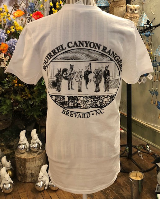 A white short-sleeved crew neck t-shirt with 'Squirrel Canyon Rangers' text and an illustration of a band on the back, displayed in a retail setting with toys and decorations in the background.