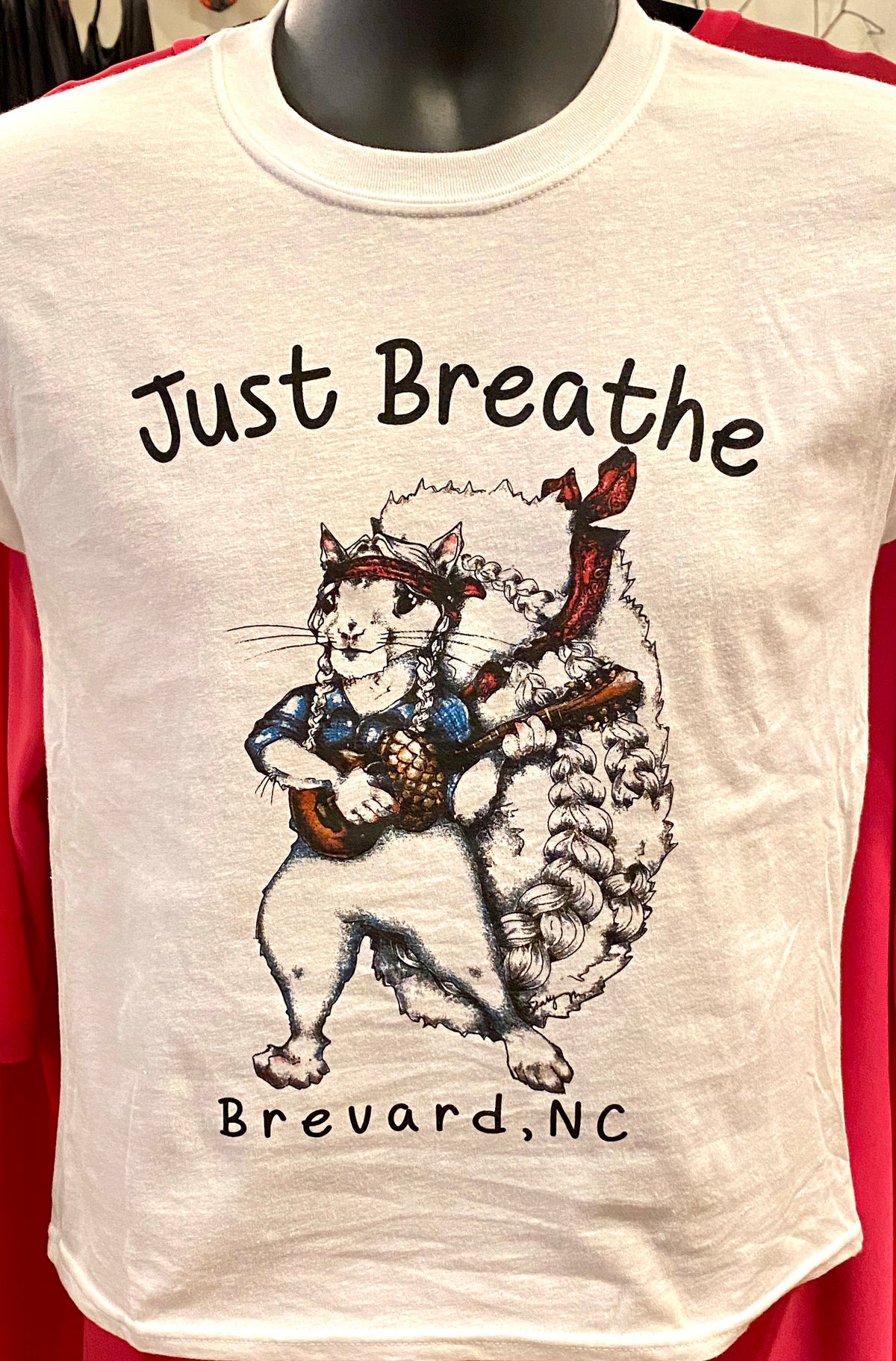 Close up view of A white youth T shirt with a Willie Nelson white squirrel graphic with the words Just Breathe and Brevard, NC