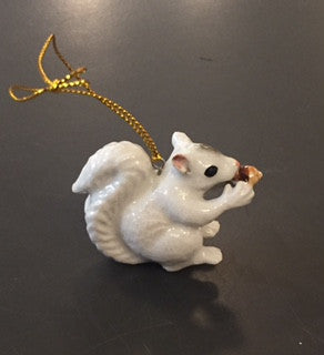 A hand painted  porcelain white squirrel ornament with a gold hanging loop, holding an acorn in its mouth.
